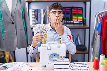 Young hispanic man dressmaker designer holding dollars covering mouth with hand, shocked and afraid for mistake. surprised expression