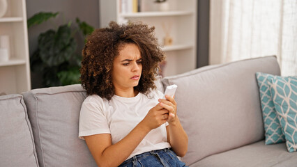 Young beautiful hispanic woman sitting on sofa using air condition remote control at home