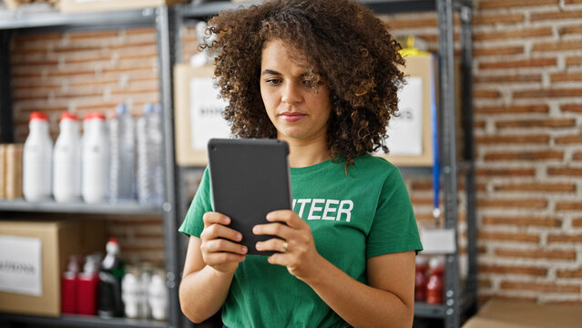 Young beautiful hispanic woman volunteer using touchpad with serious face at charity center