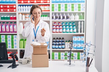Young blonde woman pharmacist holding pills bottle talking on smartphone at pharmacy