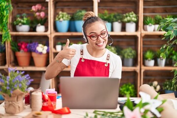 Young hispanic woman working at florist shop doing video call smiling happy and positive, thumb up doing excellent and approval sign
