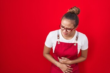 Young hispanic woman wearing waitress apron over red background with hand on stomach because...