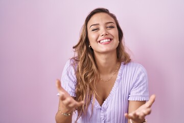 Young hispanic woman standing over pink background smiling cheerful offering hands giving assistance and acceptance.