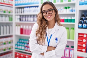 Young beautiful hispanic woman pharmacist smiling confident standing with arms crossed gesture at pharmacy - obrazy, fototapety, plakaty
