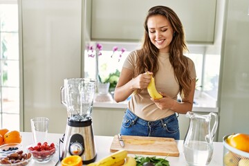 Young beautiful hispanic woman preparing vegetable smoothie with blender peeling banana at the...