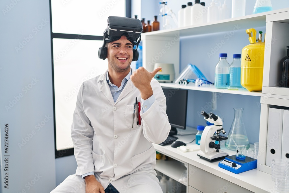 Sticker Young hispanic man working at scientist laboratory wearing vr glasses pointing thumb up to the side smiling happy with open mouth