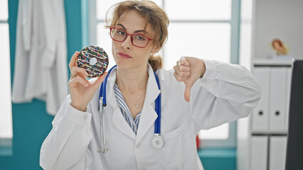 Young woman doctor holding doughnut doing thumb down gesture at the clinic