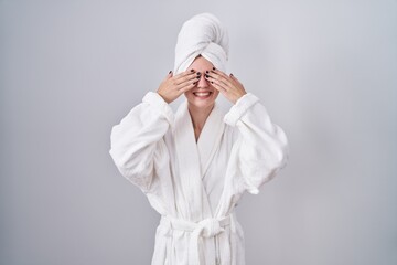 Blonde caucasian woman wearing bathrobe covering eyes with hands smiling cheerful and funny. blind concept.