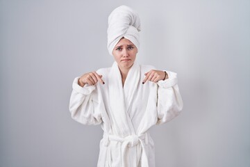 Blonde caucasian woman wearing bathrobe pointing down looking sad and upset, indicating direction with fingers, unhappy and depressed.