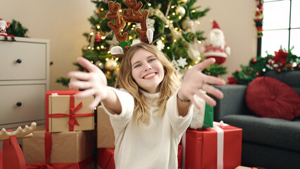 Young blonde woman sitting on floor by christmas tree with arms open at home