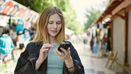 Young blonde woman using smartphone smiling at street market