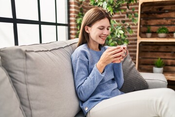 Young blonde woman drinking coffee sitting on sofa at home