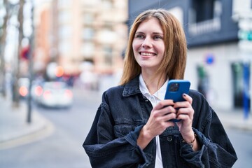 Young blonde woman using smartphone smiling at street