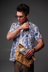 Trendy brunette man in sunglasses and aloha shirt holding money and paper bag isolated on grey
