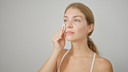 Young blonde woman cleaning face with cotton pad over isolated white background