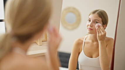 Young blonde woman cleaning face with cotton pad looking on mirror at home