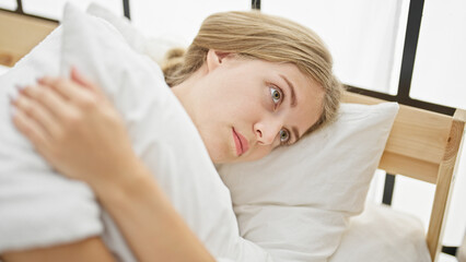 Fototapeta na wymiar Young blonde woman wearing bathrobe hugging pillow with serious face at bedroom
