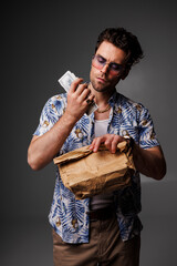 Fashionable man in 90s outfit holding dollars and paper bag on grey background, fashion concept