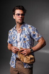 Stylish man in sunglasses and aloha shirt holding paper bag with dollars on grey 