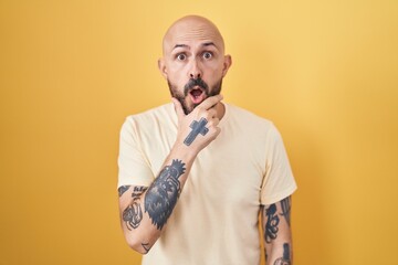 Hispanic man with tattoos standing over yellow background looking fascinated with disbelief, surprise and amazed expression with hands on chin