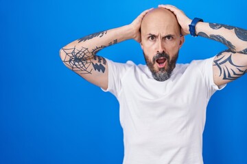 Naklejka premium Hispanic man with tattoos standing over blue background crazy and scared with hands on head, afraid and surprised of shock with open mouth