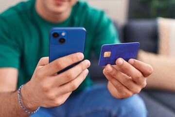 Young hispanic man using smartphone and credit card sitting on sofa at home