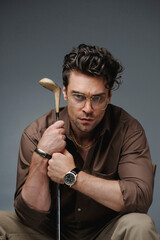 Fashionable man in luxury accessories and shirt holding golf club and looking at camera isolated on grey 