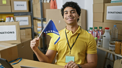 Young latin man volunteer holding bosnia and herzegovina flag smiling at charity center