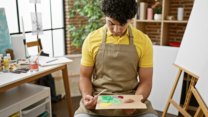 Young latin man artist holding paintbrush and palette sitting with serious face at art studio