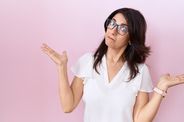 Middle age hispanic woman wearing casual white t shirt and glasses clueless and confused with open arms, no idea concept. - obrazy, fototapety, plakaty
