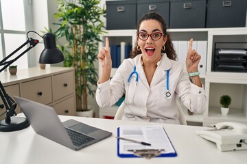 Young hispanic woman wearing doctor uniform and stethoscope smiling amazed and surprised and pointing up with fingers and raised arms.