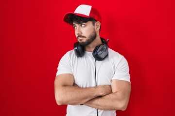 Hispanic man with beard wearing gamer hat and headphones looking to the side with arms crossed convinced and confident