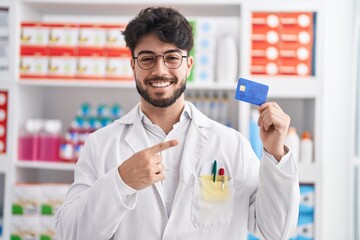 Hispanic man with beard working at pharmacy drugstore holding credit card smiling happy pointing with hand and finger