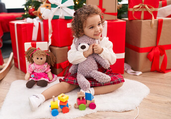 Fototapeta na wymiar Adorable hispanic toddler hugging teddy bear sitting on floor by christmas gifts at home