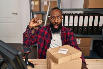 African american man working at small business ecommerce holding bitcoin scared and amazed with open mouth for surprise, disbelief face