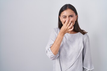 Young hispanic woman standing over white background smelling something stinky and disgusting,...