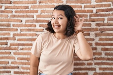 Young hispanic woman standing over bricks wall smiling with hand over ear listening an hearing to rumor or gossip. deafness concept.