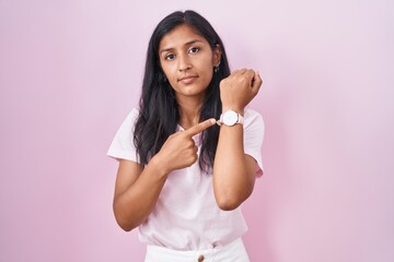 Young hispanic woman standing over pink background in hurry pointing to watch time, impatience,...