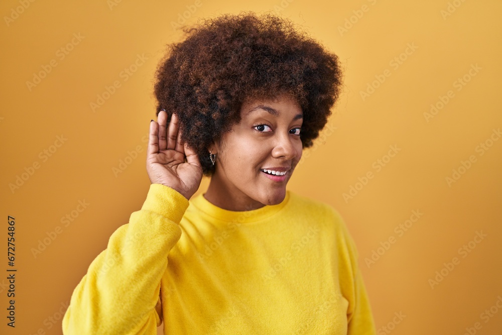 Wall mural Young african american woman standing over yellow background smiling with hand over ear listening an hearing to rumor or gossip. deafness concept.