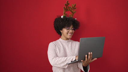 Young african american woman wearing christmas reindeer ears using laptop over isolated red...