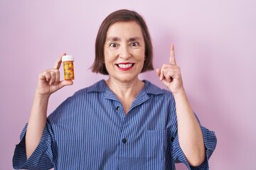 Middle age hispanic woman holding pills surprised with an idea or question pointing finger with happy face, number one