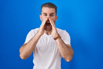 Young caucasian man standing over blue background shouting angry out loud with hands over mouth