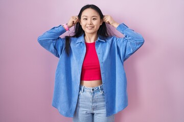 Young asian woman standing over pink background smiling pulling ears with fingers, funny gesture....