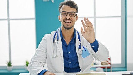 Young hispanic man doctor smiling confident saying hello with hand at clinic