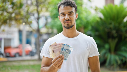 Young hispanic man holding mexican pesos at park - obrazy, fototapety, plakaty