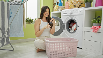 Young beautiful hispanic woman washing clothes using smartphone at laundry room