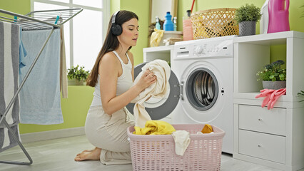 Young beautiful hispanic woman listening to music washing clothes at laundry room