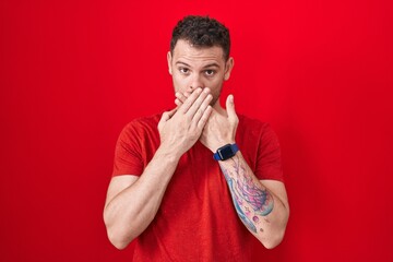 Young hispanic man standing over red background shocked covering mouth with hands for mistake....
