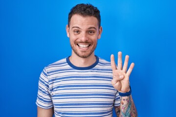 Naklejka premium Young hispanic man standing over blue background showing and pointing up with fingers number four while smiling confident and happy.