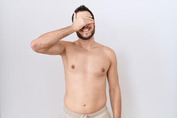 Young hispanic man standing shirtless over white background smiling and laughing with hand on face covering eyes for surprise. blind concept.